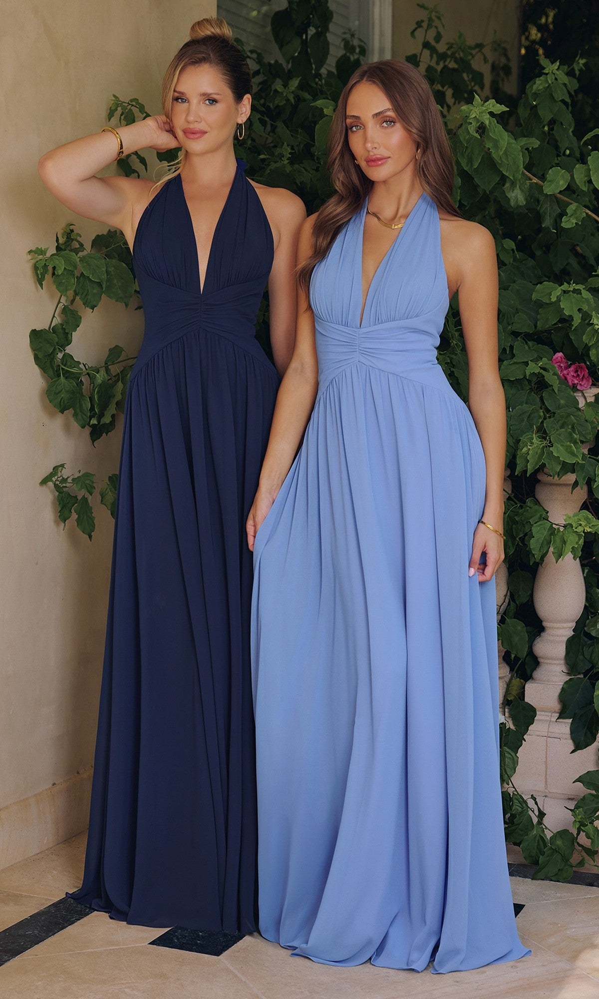 Two women pose gracefully by leafy green plants, wearing Amoris T715 long chiffon halter prom dresses—one in dark blue and the other in light blue.